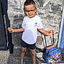 blue_crocs, boy, casual, child, glasses, luggage, outdoor, paw_patrol, person, playful, shorts, sidewalk, smiling, snack, standing, stone_wall, suitcase, sunny, white_tshirt, young
