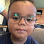 blue_shirt, brick_wall, candid, casual_clothing, child, closeup, curly_hair, expressionless, eyewear, face, furniture, glasses, head_and_shoulders, indoor, modern_interior, person, portrait, seated, television, young_child