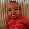 baby, infant, tongue_out, red_clothing, smiling, face, head, eyes, skin, cheeks, seated, indoors, ribbed_background, cute, young_child, portrait, happy, person, baby_girl, cozy