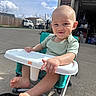 baby, child, high_chair, outdoor, sunny, sky, clouds, pavement, smile, pointing, barefoot, green_clothing, toy, garage, fence, vehicle, seat, person, happy, daytime