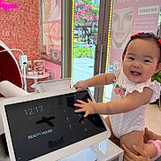 Aliya is registered to the contest to win money with this photo: baby, child, smiling, touchscreen, tablet, beauty_salon, flower_wall, indoor, red_bows, white_onesie, hands, adult_hand, counter, screen, digital_display, window, daylight, happy, cute, technology
