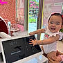 baby, child, smiling, touchscreen, tablet, beauty_salon, flower_wall, indoor, red_bows, white_onesie, hands, adult_hand, counter, screen, digital_display, window, daylight, happy, cute, technology