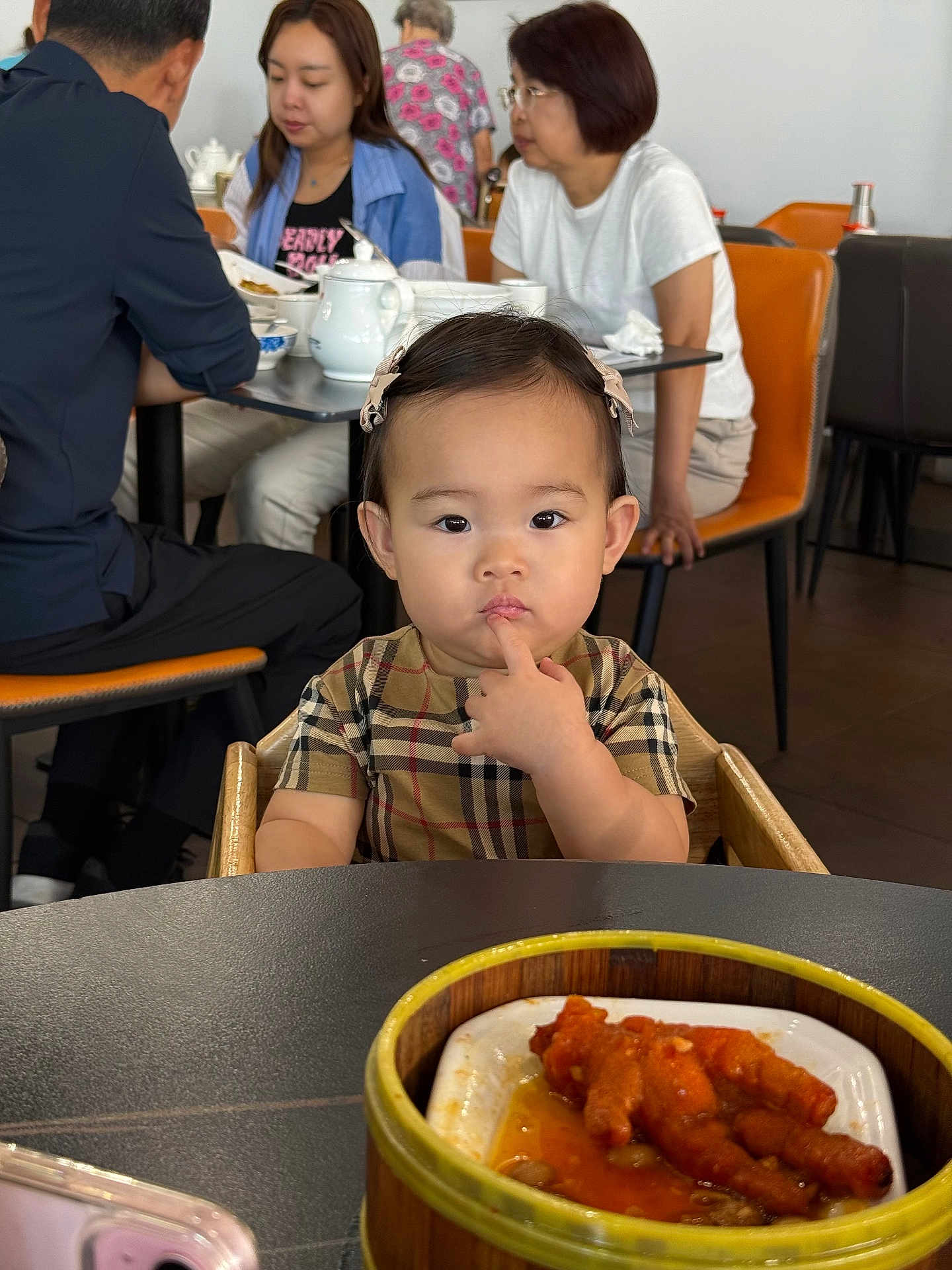 Aliya joined the competition — help win amazing prizes! toddler, child, high_chair, plaid_shirt, hair_bows, restaurant, table, food, chicken_feet, asian, people, dining, adult, seated, furniture, cup, teapot, plate, curious, indoor