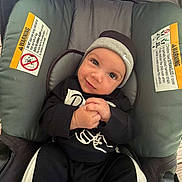 Azael is registered to the contest to win money with this photo: baby, black_outfit, carrier, child, clothing, comfortable, cushion, cute, face, hands_clasped, hat, headwear, indoor, infant, person, portrait, seat, smile, warning_label, young