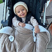Kareem is registered to the contest to win money with this photo: baby, child, smiling, blanket, stroller, hat, outdoor, cozy, cute, happy, person, infant, warm, clothing, seated, playful, face, babywearing, bundle, headwear