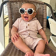 Kareem is registered to the contest to win money with this photo: baby, child, sunglasses, chair, wicker, smiling, drool, outdoor, porch, clothing, feet, happy, infant, cute, casual, person, seat, relaxed, sunlight, fashion