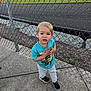 Luca joined the competition — help win amazing prizes! toddler, child, boy, standing, sidewalk, chain_link_fence, outdoor, concrete, clothing, sneakers, short_sleeve_shirt, casual, daylight, curious_expression, blonde_hair, young_child, graphic_tshirt, pavement, sunlight, nature_background