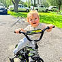 Luca is registered to the contest to win money with this photo: toddler, child, bike, bicycle, training_wheels, outdoor, sunlight, driveway, grass, tree, car, smiling, happy, daytime, casual_clothing, play, young_child, person, nature, recreation