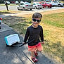 child, boy, sunglasses, cooler, wheeled_cooler, summer, outdoor, sidewalk, grass, shadow, casual_clothing, shorts, long_sleeve_shirt, footwear, parking_lot, cars, trees, sunny, smile, walking