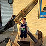 child, boy, smiling, tank_top, dinosaur_print, anchor, rusty, chain, wood_post, concrete_floor, brick_wall, outdoor, sunlight, shadow, nautical, sign, casual_clothing, happy, summer, daytime