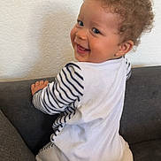 Leeroy a rejoint le concours — aidez-le/la à gagner de superbes lots ! baby, child, curly_hair, smiling, white_outfit, striped_clothing, couch, indoor, happy, sitting, person, face, cute, infant, young_child, playful, home, furniture, portrait, looking_back
