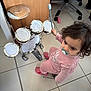 child, toddler, pacifier, toy_drum, drumstick, musical_instrument, tile_floor, office_chair, pink_outfit, slippers, curly_hair, indoor, playtime, looking_up, facial_expression, standing, wooden_cabinet, building_blocks, small_drum_set, floor_tiles