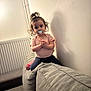 child, toddler, pacifier, sofa, couch, living_room, wall, radiator, top_knot, curly_hair, pink_shirt, navy_leggings, pink_shoe, sitting, climbing, curious_expression, big_eyes, shadow, indoor, furniture