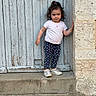 toddler, child, standing, door, wooden_door, stone_step, outdoor, curly_hair, white_tshirt, heart_pattern, leggings, sneakers, concrete, wall, playful, casual_clothing, young_child, person, portrait, expression