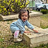 child, toddler, girl, denim_dress, pigtails, outdoor, gravel, concrete_block, playing, smiling, happy, greenery, plants, shoes, white_tights, parked_cars, daylight, curly_hair, nature, crouching