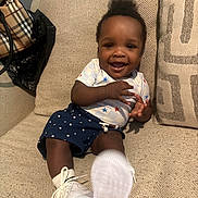 Dakari is registered to the contest to win money with this photo: baby, child, smiling, sitting, couch, white_shoes, star_pattern, clothing, happy, indoor, foot, person, cute, infant, young_child, textile, furniture, sock, hand, portrait