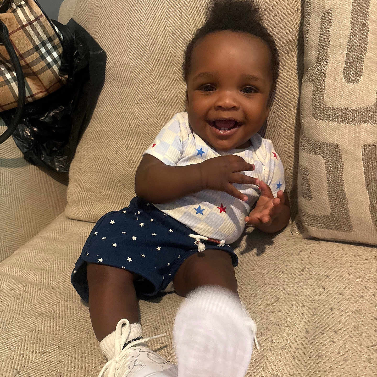 Dakari is registered to the contest to win money with this photo: baby, child, smiling, sitting, couch, white_shoes, star_pattern, clothing, happy, indoor, foot, person, cute, infant, young_child, textile, furniture, sock, hand, portrait