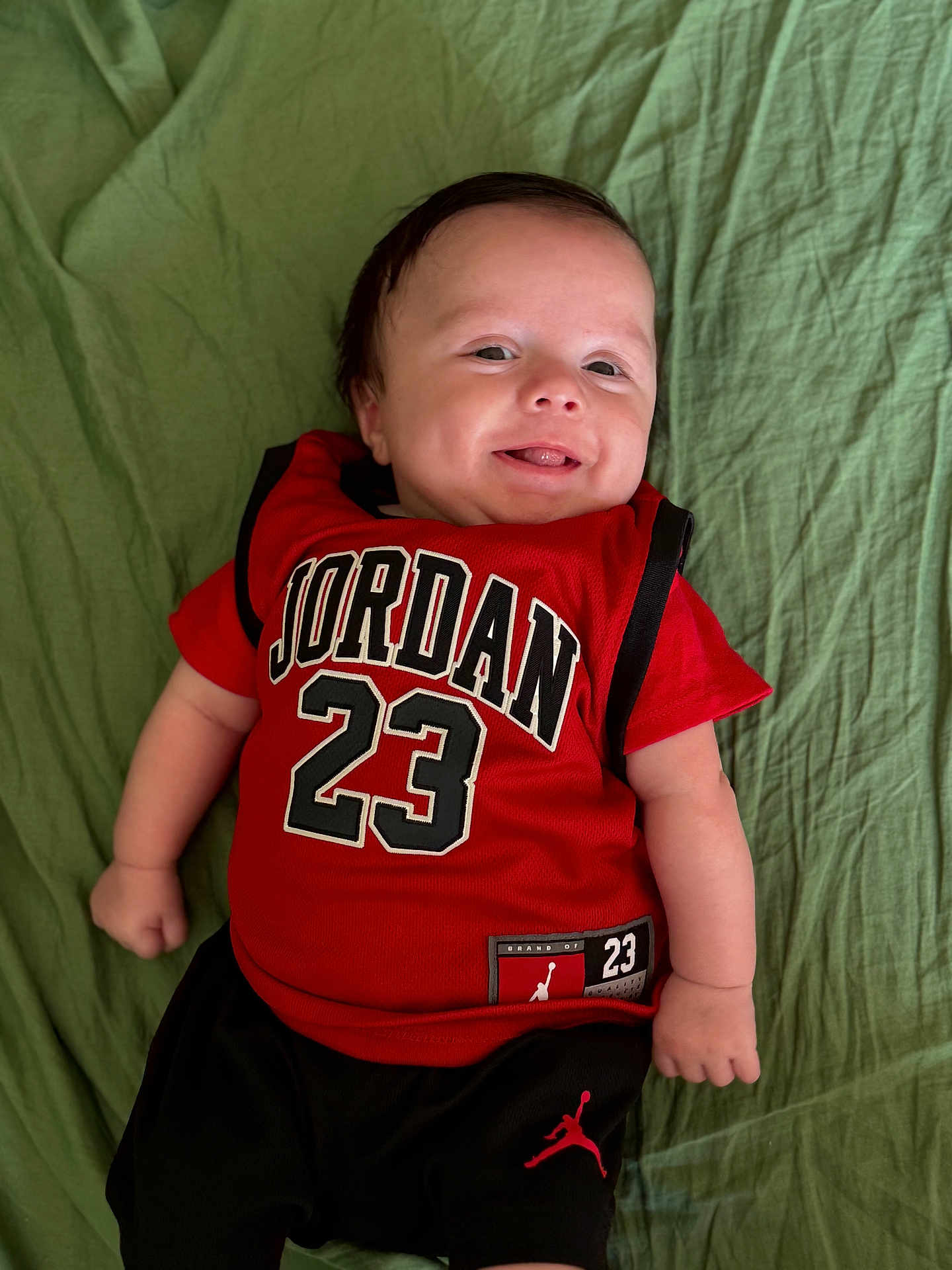 Giulian a rejoint le concours — aidez-le/la à gagner de superbes lots ! baby, smiling, infant, clothing, red_shirt, black_shorts, green_blanket, portrait, happy, cute, child, person, young, indoors, casual, resting, lying_down, short_sleeves, sportswear, jordan
