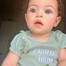 toddler, child, curly_hair, blue_eyes, indoor, shirt, green_clothing, pineapple_design, baby, portrait, casual, cute, person, young_child, soft_light, expression, sitting, real_photo, face, curiosity