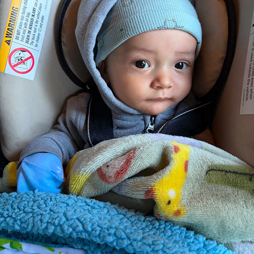 Zaiel is registered to the contest to win money with this photo: baby, blanket, blue, car_seat, child, clothing, comfort, cozy, cute, expression, face, hat, indoor, infant, looking, person, portrait, seatbelt, warm, young