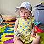 toddler, child, hat, dress, toy, floor_mat, indoor, sitting, colorful, play_area, baby, foot, hand, face, puzzle_mat, yellow, white, blue, flower_pattern, soft_light