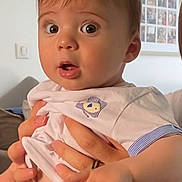 Liam a rejoint le concours — aidez-le/la à gagner de superbes lots ! baby, child, face, hand, shirt, shorts, person, indoor, surprised, expression, skin, adult_hand, watch, photo_wall, home, furniture, sitting, holding, clothing, portrait