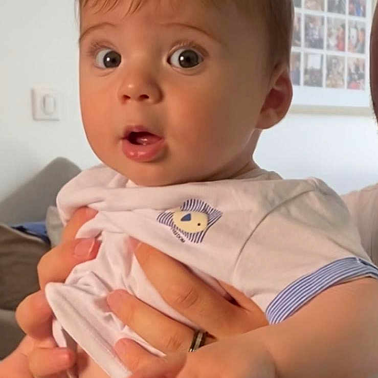 Liam a rejoint le concours — aidez-le/la à gagner de superbes lots ! adult_hand, baby, child, clothing, expression, face, furniture, hand, holding, home, indoor, person, photo_wall, portrait, shirt, shorts, sitting, skin, surprised, watch