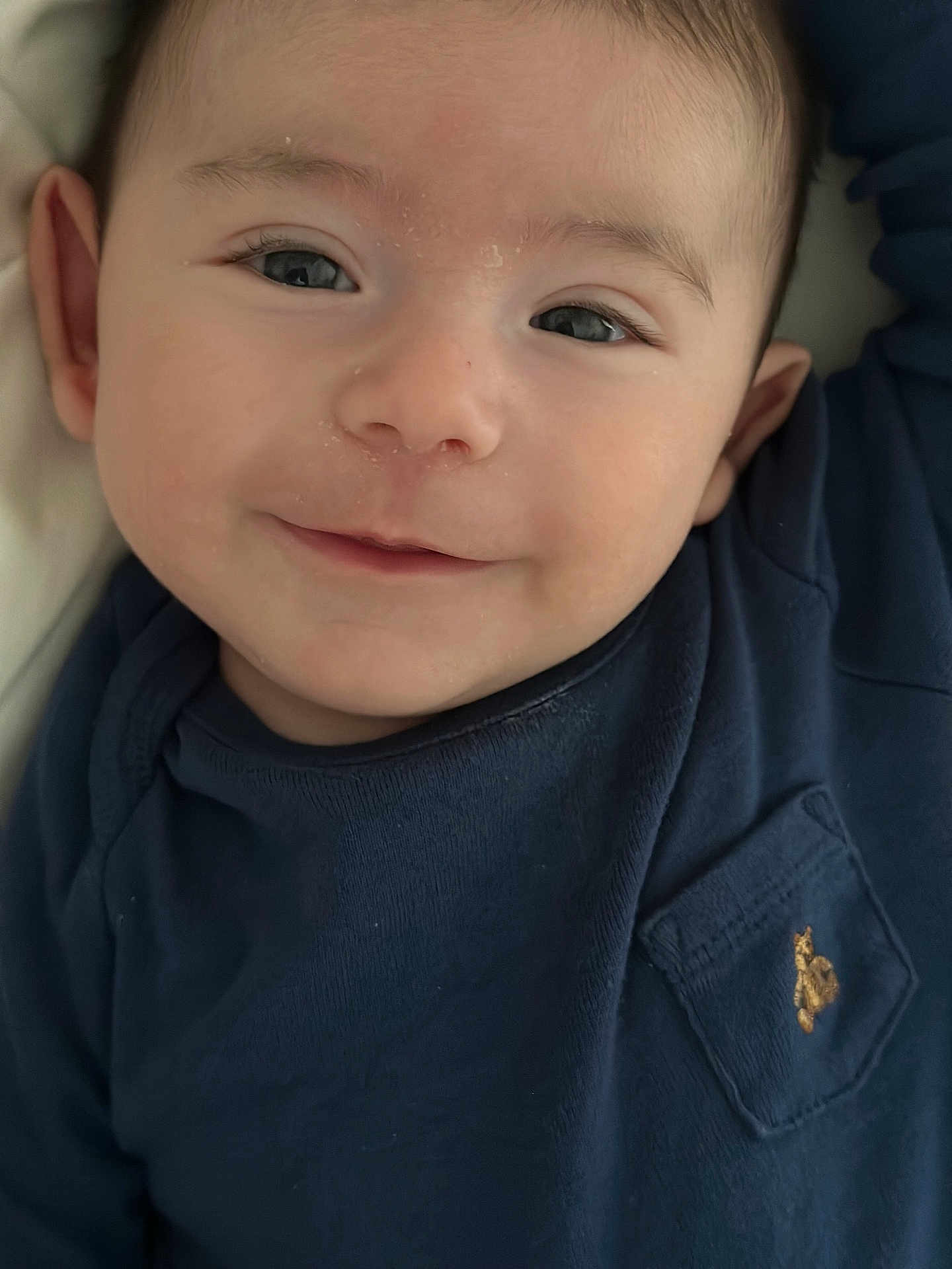 James is registered to the contest to win money with this photo: baby, smile, face, close_up, child, infant, blue_shirt, pocket, embroidery, skin, eyes, head, person, portrait, cute, young, happy, indoors, clothing, baby_face