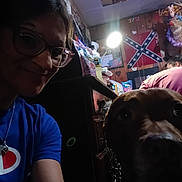 Doobie joined the competition — help win amazing prizes! person, dog, glasses, blue_shirt, indoor, lamp, snacks, wall_decorations, confederate_flag, wood_paneling, furniture, chain_collar, face, smile, close_up, head, brown_dog, clutter, casual, evening