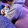 baby, child, person, hoodie, couch, blanket, indoor, carpet, purple, seated, holding, young_person, casual_clothing, curly_hair, comfort, relaxed, home, infant, light, portrait