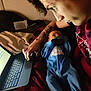 adult, baby, bed, blue_onesie, computer, digital_device, face, hand, indoor, laptop, person, tattoo, technology, typing, lying_down, pacifier, blanket, clothing, bedroom, focus