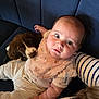baby, bib, blue_couch, brown_and_white_dog, child, close_up, couch, curious_eyes, cute, cute_expression, floral_pattern, indoor, infant, pet, puppy, relaxed, snuggling, soft_lighting, striped_sleeve, textured_pants