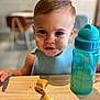 Loan a rejoint le concours — aidez-le/la à gagner de superbes lots ! toddler, child, face, eyes, messy_face, cookie, broken_cookie, table, sippy_cup, drink, blue, indoor, blurred_background, wooden_table, expression, curious, snack, young_child, sitting, bib
