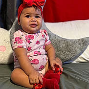 Mía joined the competition — help win amazing prizes! baby, child, red_bow, headband, floral_onesie, red_socks, bed, pillow, gray_pillow, white_pillow, red_background, cute, smiling, indoors, portrait, sitting, infant, person, cozy, home