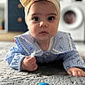 baby, child, face, headband, bow, blue_clothing, patterned_clothing, hand, floor, gray_rug, indoor, home, washing_machine, background_blur, curious, eyes, portrait, infant, crawling, closeup