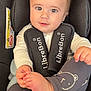 baby, child, car_seat, infant, face, eyes, clothing, white_shirt, leggings, bear_pattern, foot, hand, hair, seat_belt, black, safety, cute, portrait, indoor, closeup