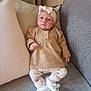 baby, infant, couch, pillow, headband, slippers, bear_slippers, clothing, leggings, sitting, indoor, portrait, cute, expression, face, small_child, soft_furniture, cozy, neutral_colors, child