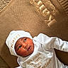 newborn, baby, infant, child, portrait, lying_down, white_outfit, white_hat, couch, upholstery, indoor, face, eyes, mouth, clothing, embroidery, text_on_clothing, brown_background, soft_lighting, photography