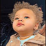 child, curly_hair, blue_eyes, beige_jacket, zipper, portrait, close_up, face, cute, baby, earrings, looking_up, indoor, dark_background, heart_graphic, casual_clothing, young_child, adorable, skin, fashion
