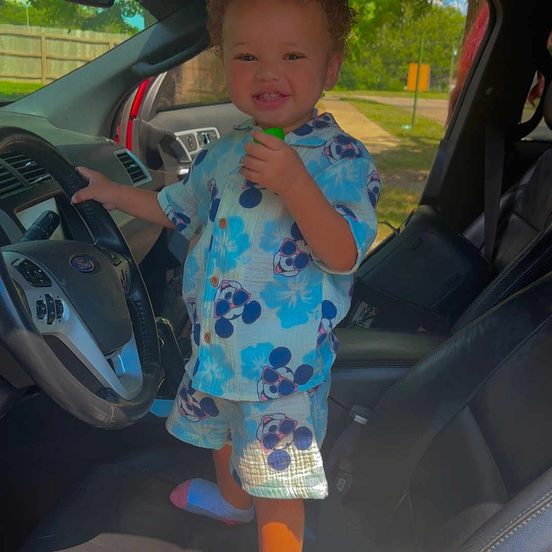 Deair is registered to the contest to win money with this photo: car, child, clothing, curly_hair, green_object, happy, interior, leather_seat, mickey_mouse, playful, seat, shirt, shorts, smiling, socks, standing, steering_wheel, sunlight, toddler, window