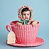 baby, child, teacup, pink, woven, hat, floral_bonnet, blue_background, cute, portrait, infant, face, hands, saucer, ribbon, bow, tag, playful, studio, photography