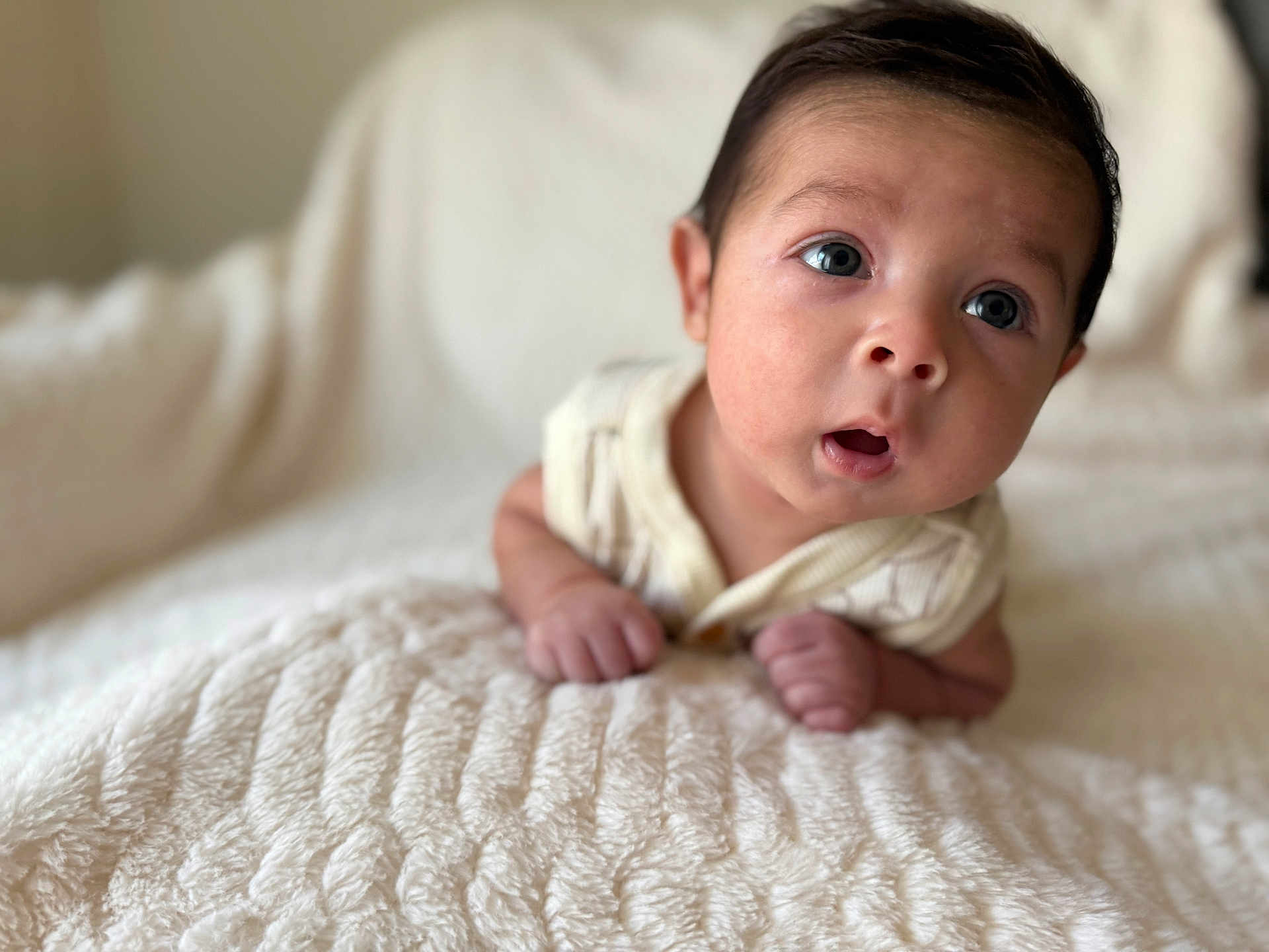 Aramis is registered to the contest to win money with this photo: baby, infant, face, eyes, blanket, soft_texture, indoor, portrait, curious, hands, expression, cute, child, young, skin, clothing, head, lying_down, closeup, natural_light