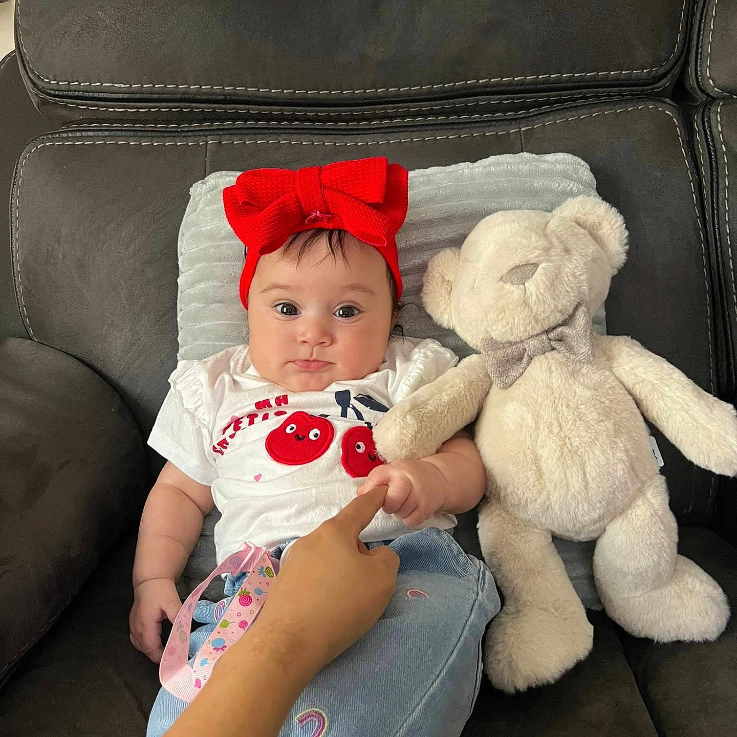 Dahlia is registered to the contest to win money with this photo: baby, child, couch, cozy, cute, finger, hand, headband, indoor, infant, jeans, person, pillow, plush_toy, rainbow_patch, red_bow, smiling, soft_toy, teddy_bear, white_shirt