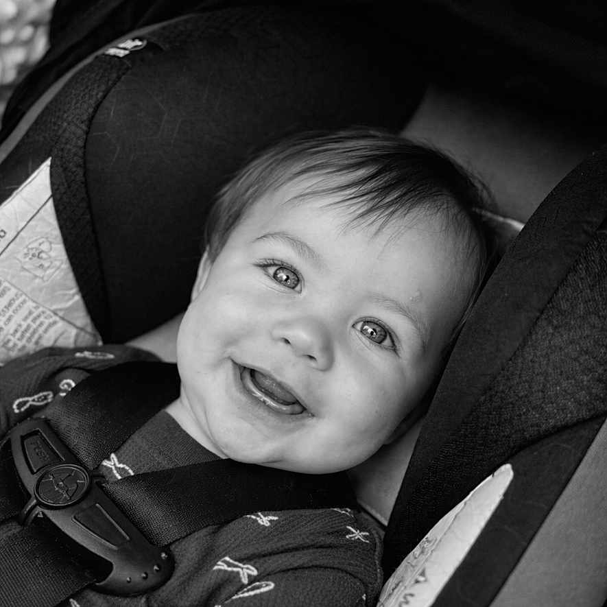 Noah is registered to the contest to win money with this photo: baby, child, car_seat, smile, face, black_and_white, infant, seatbelt, clothing, happy, portrait, safety, cute, young_child, hair, eyes, car, infancy, seat, person
