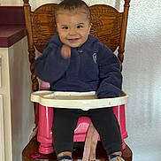 Liam joined the competition — help win amazing prizes! toddler, child, smiling, sitting, booster_seat, wooden_chair, blue_jacket, sneakers, indoor, wall, happy, person, face, hair_tied, footwear, clothing, seatbelt, table, hand, expression