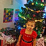 child, girl, christmas_tree, christmas_lights, christmas_decorations, presents, gift_bags, red_dress, holiday, smiling, indoor, carpet, wall, stocking, poster, alphabet_poster, number_poster, black_shoes, festive, happy