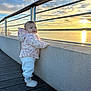 Lia participe au concours pour gagner de l'argent avec cette photo : baby, child, sunset, pier, water, sky, clouds, railing, jacket, pants, boots, outdoor, smiling, happy, nature, evening, scenic, portrait, person, winter_clothing