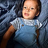 toddler, child, blue_overalls, striped_shirt, curly_hair, blue_blanket, indoor, portrait, cute, soft_texture, relaxed, baby, smiling, young_child, person, infant, casual_clothing, laying_down, light, skin