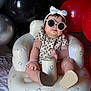 baby, infant, sunglasses, headband, bow, inflatable_chair, balloons, red_balloon, black_balloon, silver_balloon, patterned_dress, sitting, chubby_arms, rosy_cheeks, party, celebration, portrait, cute, accessory, nursery