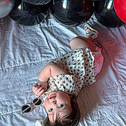 Ainoha is registered to the contest to win money with this photo: child, toddler, baby, lying_down, bed, blanket, balloons, black_balloons, red_balloon, sunglasses, headband, bow, dress, floral_pattern, hair, face, indoor, party, reflection, playful